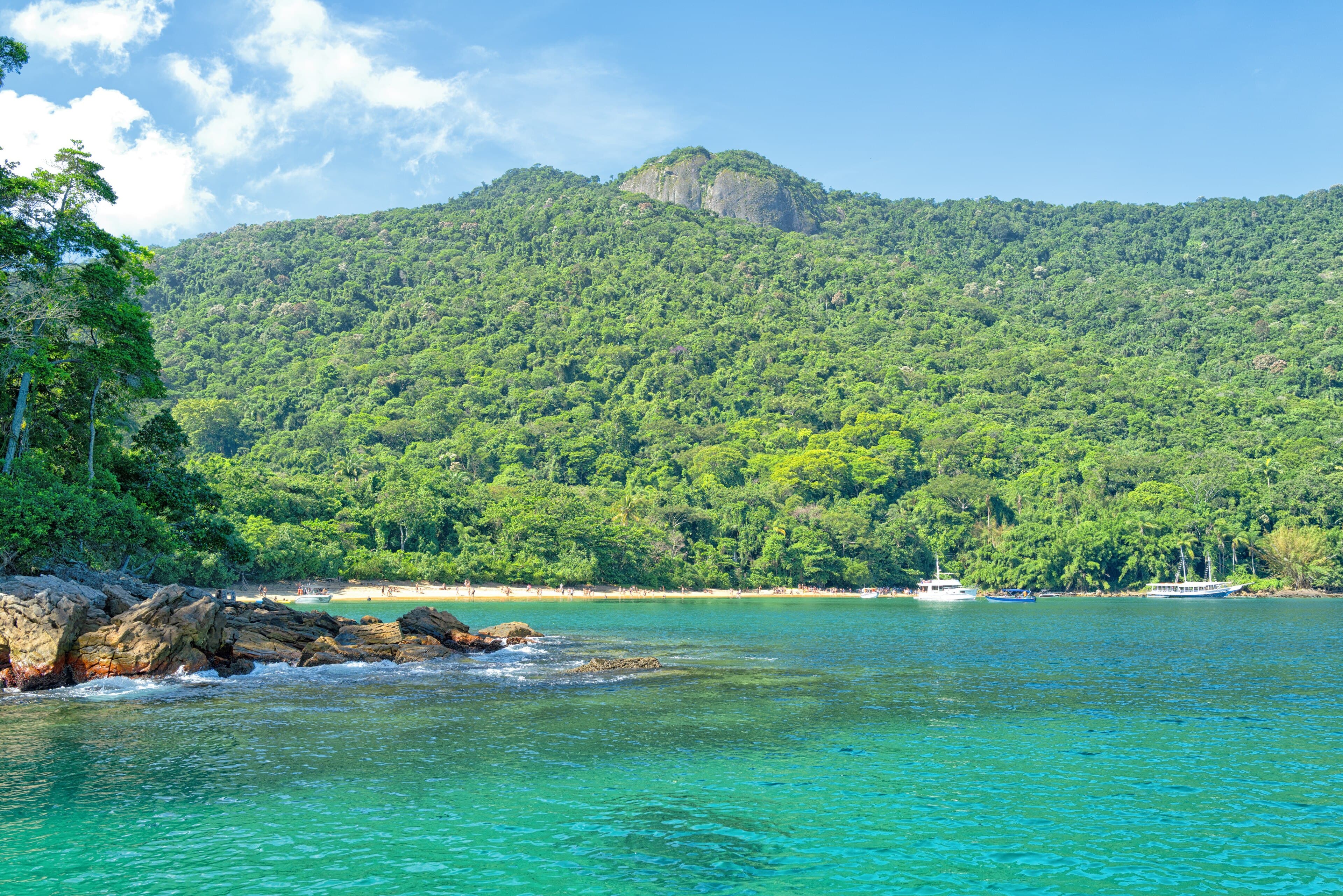 Praia com embarcações ancoradas em água esverdeada e morro coberto de mata ao fundo, na região de Angra dos Reis